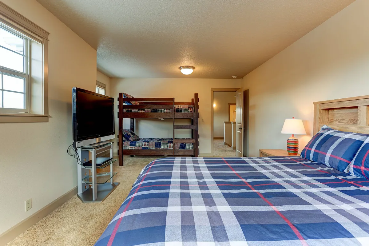 A playful family bedroom featuring bunk beds, a bed with plaid bedding, a television, and a warm lamp. The layout suggests a space where kids and adults can watch TV, play games, or enjoy sleepovers together.