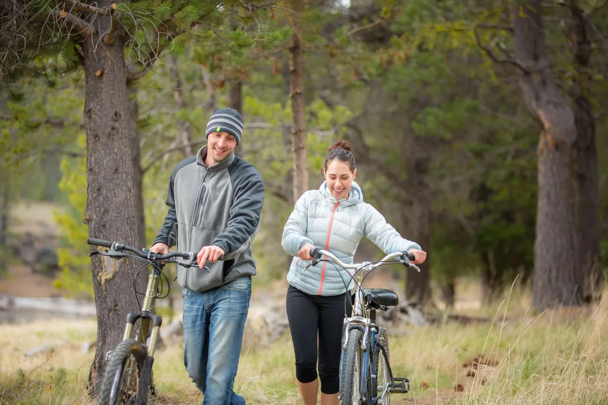 This image features two people walking with bikes along a forested trail, surrounded by trees and natural landscape. The scene captures the spirit of outdoor adventure, with the individuals appearing playful and ready to explore Central Oregon’s natural beauty.