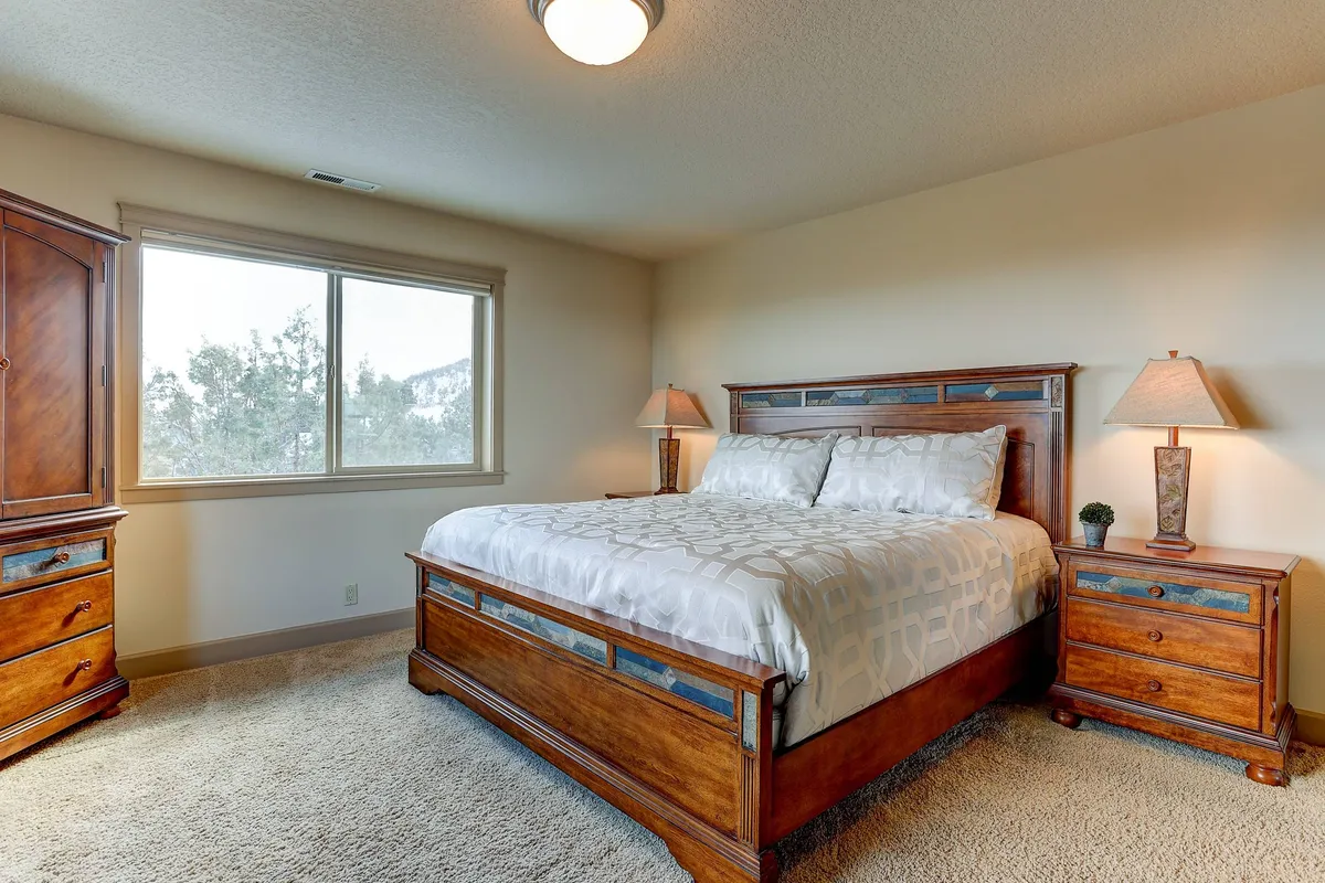 A cozy bedroom scene with a comfortable bed framed by windows, nightstands, and soft lighting from lamps. The view out the window suggests a tranquil, scenic backdrop, enhancing the atmosphere of peacefulness.