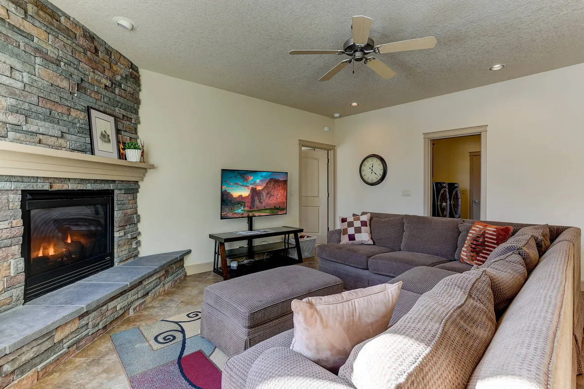 A spacious living room featuring a cozy sectional sofa facing a prominent fireplace, with decorative pillows adding a touch of warmth. A television is mounted above the mantle, and large windows fill the room with natural light, creating an inviting atmosphere ideal for relaxation, movie nights, or family conversations.