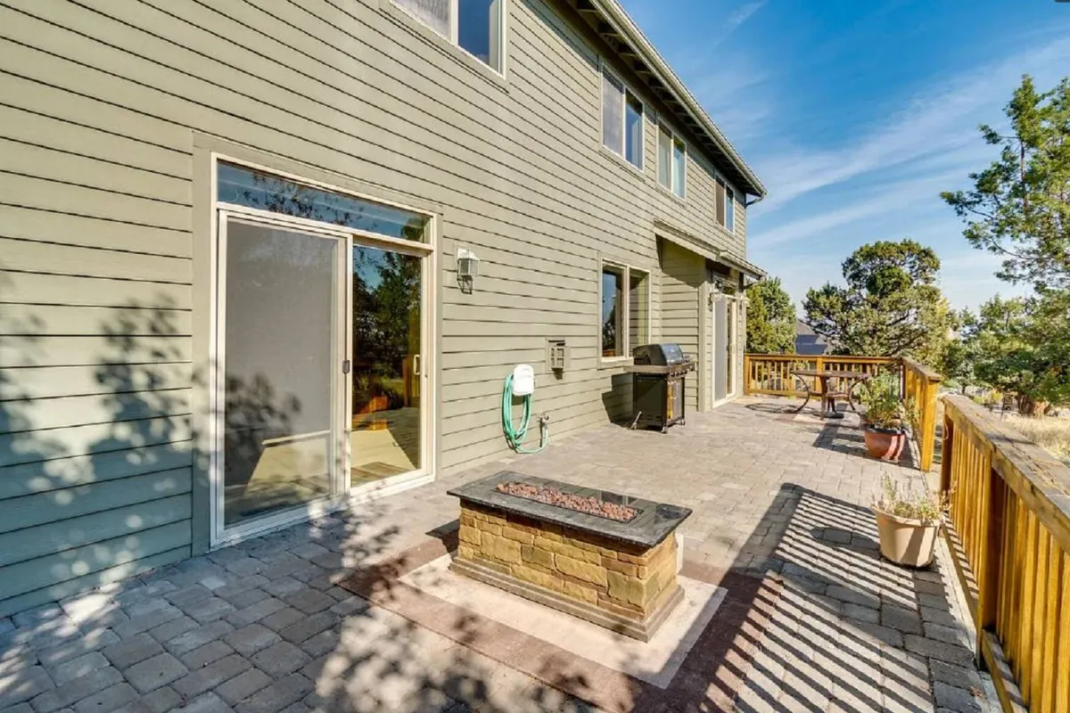 A paved patio with a fire pit, grill, and deck railing creates an atmosphere suited for gathering and enjoying the outdoors. The relaxed mood hints at sunset views, cozy evenings by the fire, and celebrations with friends or family.