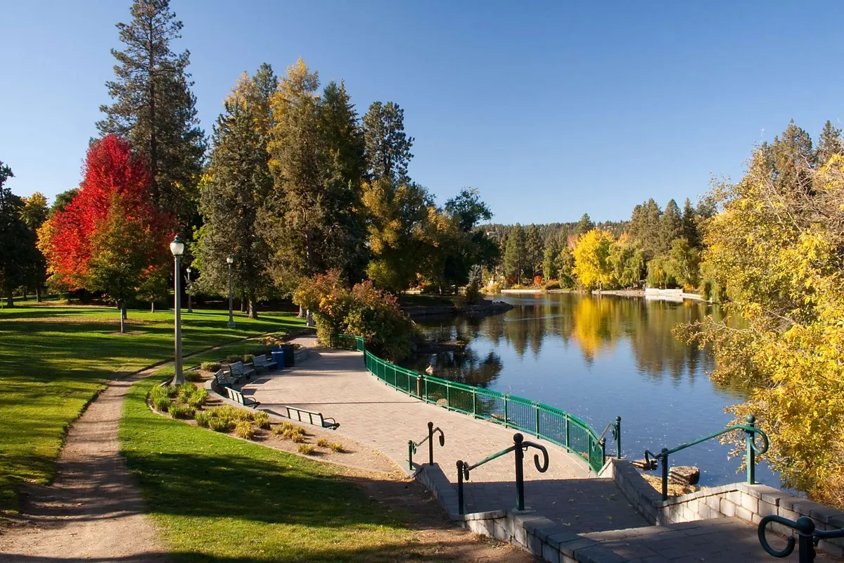 A tranquil lakeside scene featuring calm water, colorful trees, a walking path, green railing, and park benches. The area is ideal for walking, jogging, relaxing by the water, or enjoying a picnic while soaking in natural beauty.