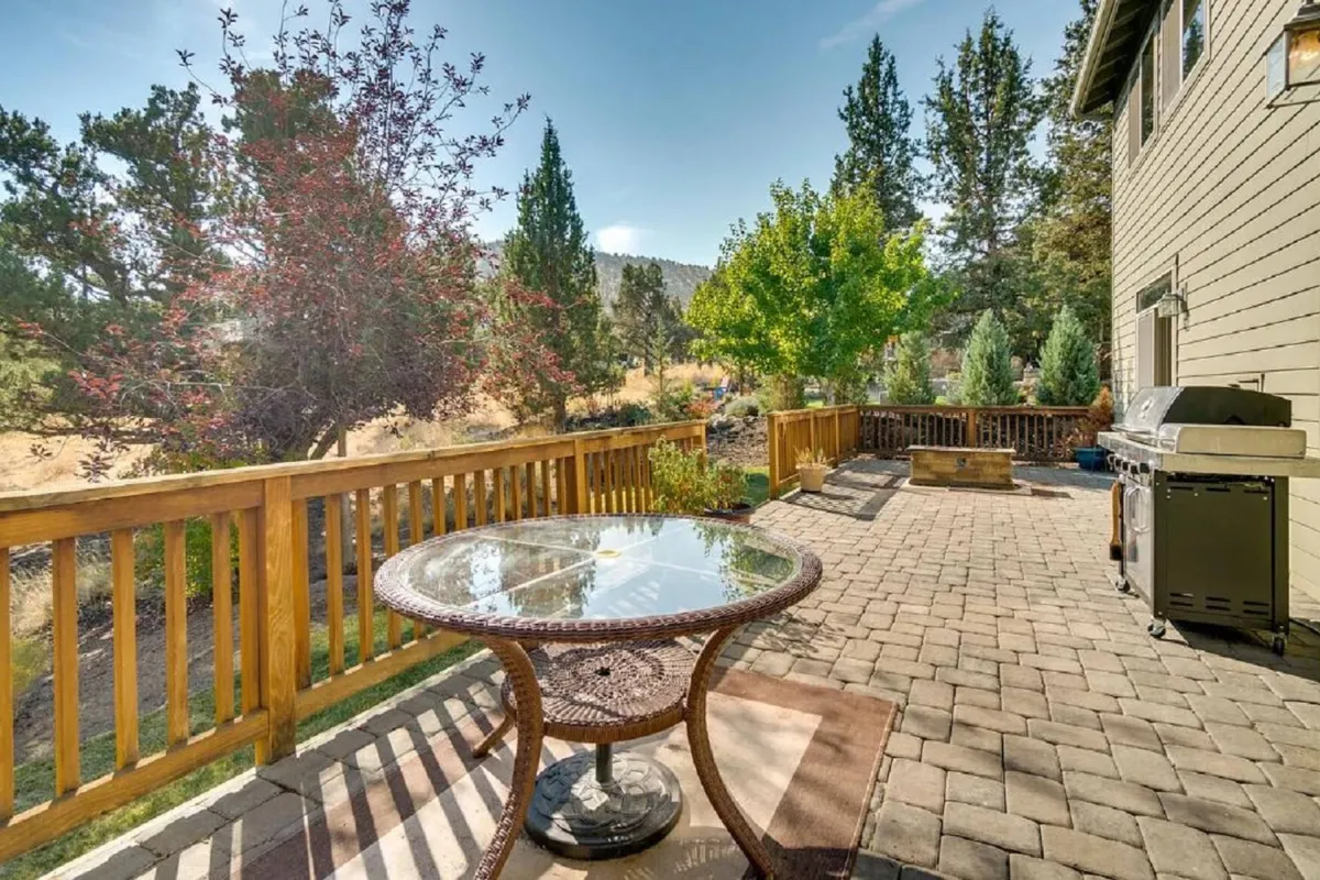 An inviting patio scene showcases a glass-top dining table, a grill ready for outdoor cooking, lush greenery, and a wooden railing. The setting radiates relaxation and social possibilities, with plenty of space for dining, barbecuing, or simply enjoying the view.