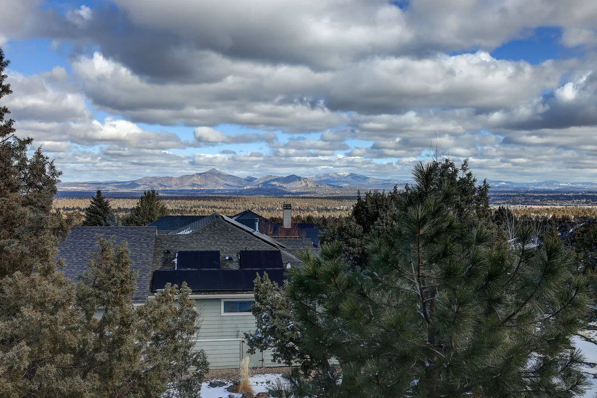 This image captures a scenic view of distant mountains, a cloudy sky, rooftops, and pine trees, evoking a sense of natural beauty and peaceful surroundings. The landscape hints at the outdoor recreational opportunities nearby and situates the property within its appealing mountain environment.