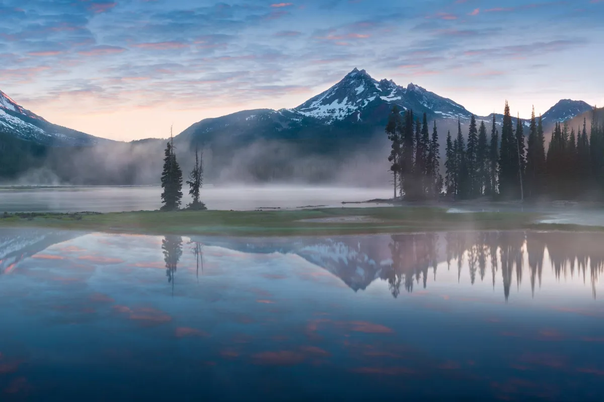 A tranquil view of mountains, a lake, and surrounding trees beneath a vast sky. The scene evokes peace and the opportunity for various outdoor activities, such as kayaking, fishing, hiking, and photography.