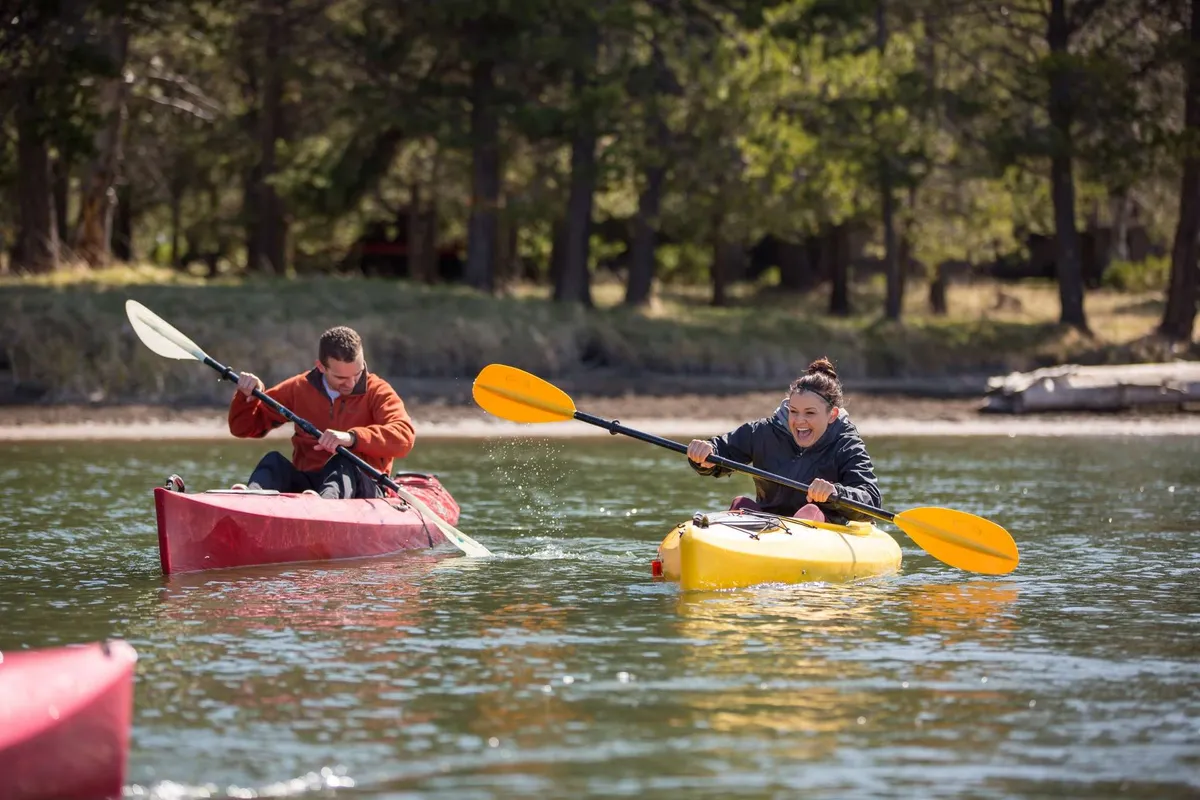 Two kayakers paddle vibrant kayaks across reflective water, capturing the spirit of playful outdoor adventure and close connection with nature. The bright colors and joyful atmosphere convey energetic fun for all ages.