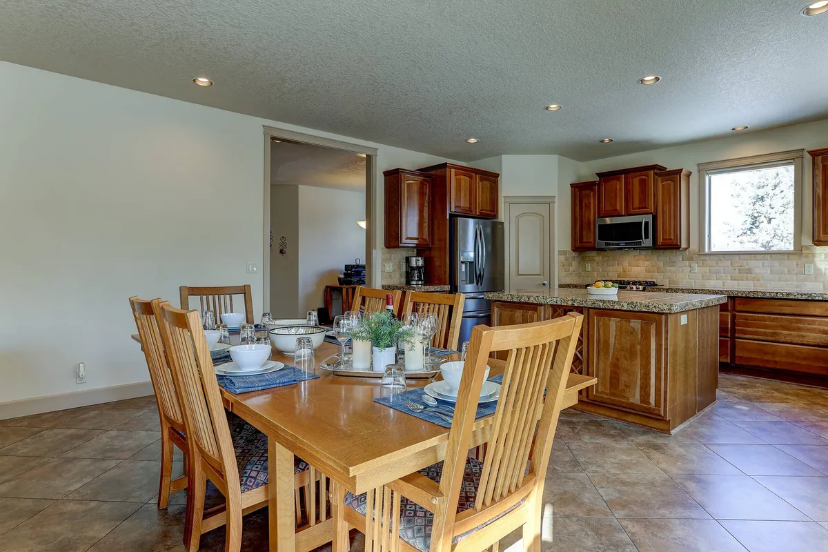 An expansive dining table is set for a meal, positioned adjacent to open kitchen cabinetry. The inviting scene suggests a space perfect for group dinners, celebrations, or lively board game nights, with plenty of natural light and a sense of openness.