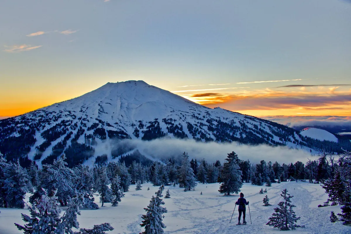 A skier moves down a snow-covered slope against a dramatic mountain peak at sunset, surrounded by snow-dusted trees. The scene is vibrant and adventurous, showcasing winter sports opportunities in a beautiful alpine environment.